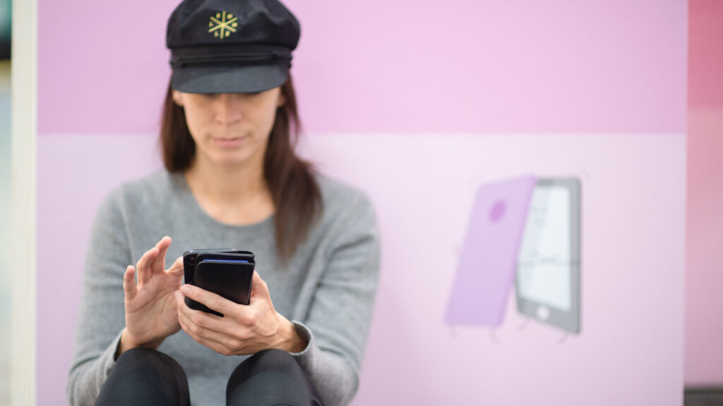 Kvinna med keps sitter framför rosa vägg och använder mobiltelefon.